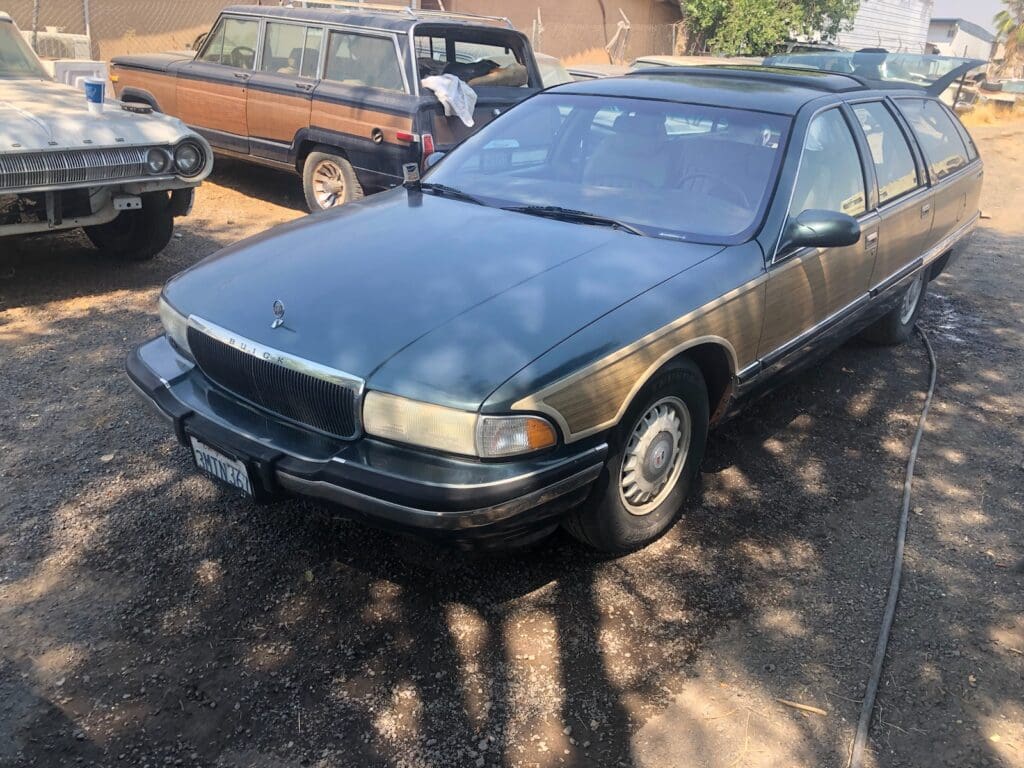 Older brown sedan parked outdoors with other vehicles nearby.