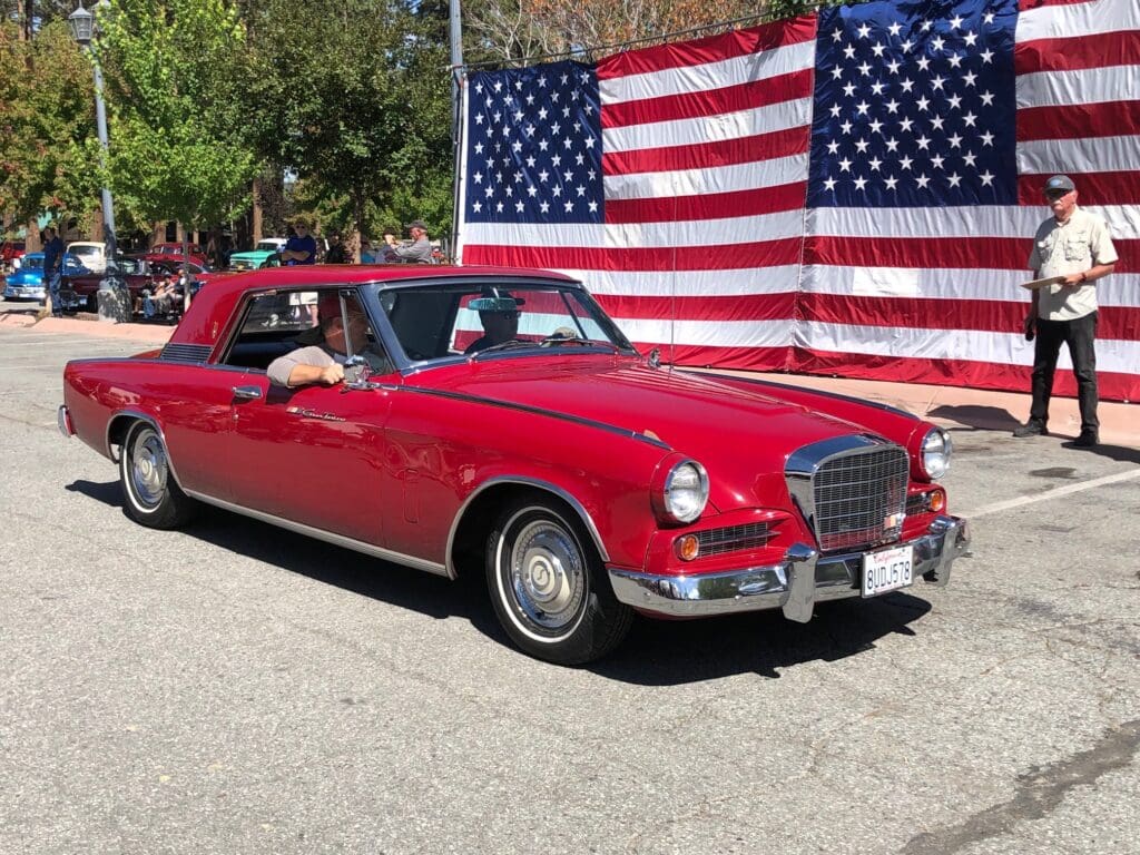 Classic red convertible car parked in front of a large American flag.