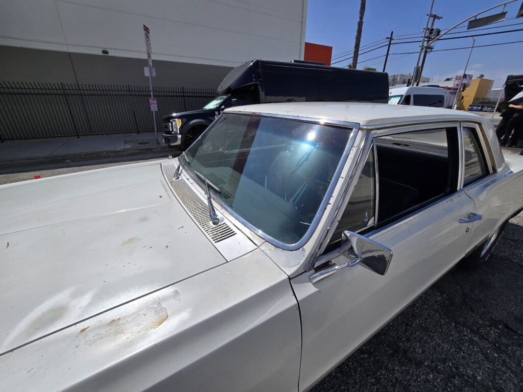 Old white car with visible rust and a black roof in a parking lot.