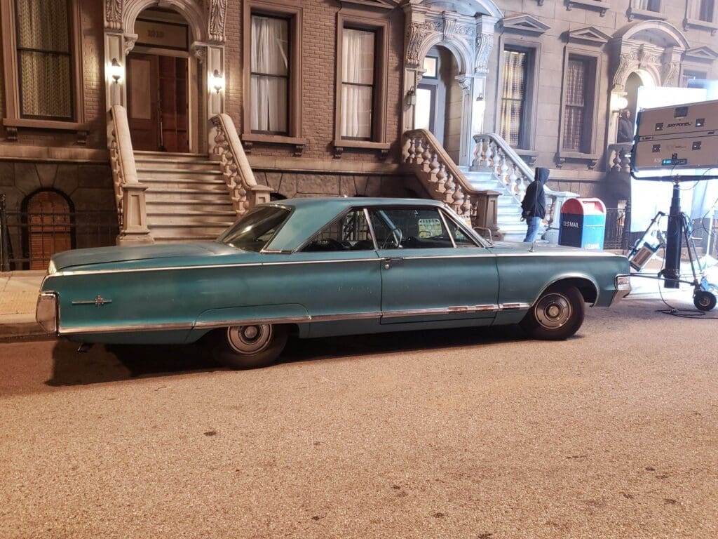 A vintage teal car parked on a city street in front of brownstone buildings.