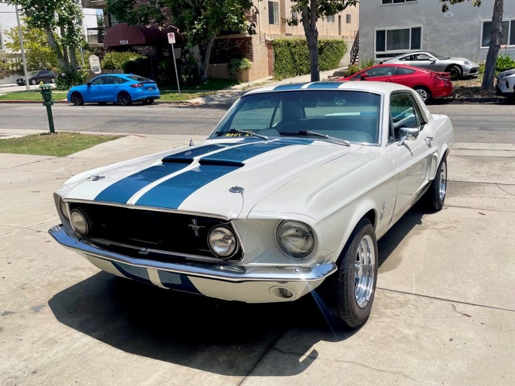 Classic white Ford Mustang with blue racing stripes parked on a sunny driveway.