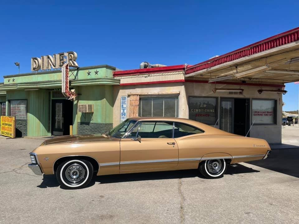 A vintage gold-colored car parked in front of retro buildings under a clear blue sky.