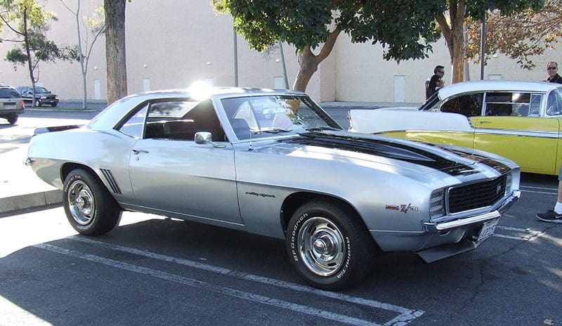 Classic silver muscle car parked under a tree in a sunny outdoor area.