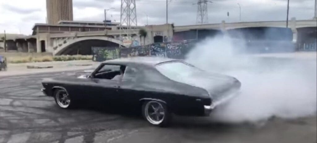 Black classic car doing a burnout with smoke around it.