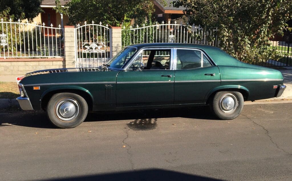 A vintage dark green car parked beside a fence on a sunny day.