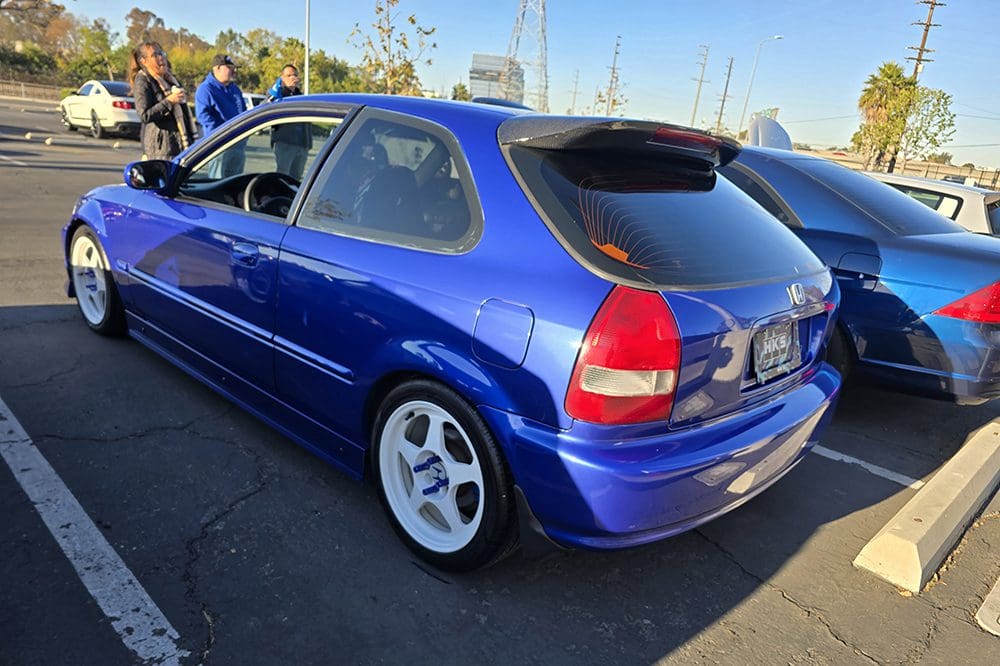 Blue hatchback car with white rims parked outdoors on a sunny day.
