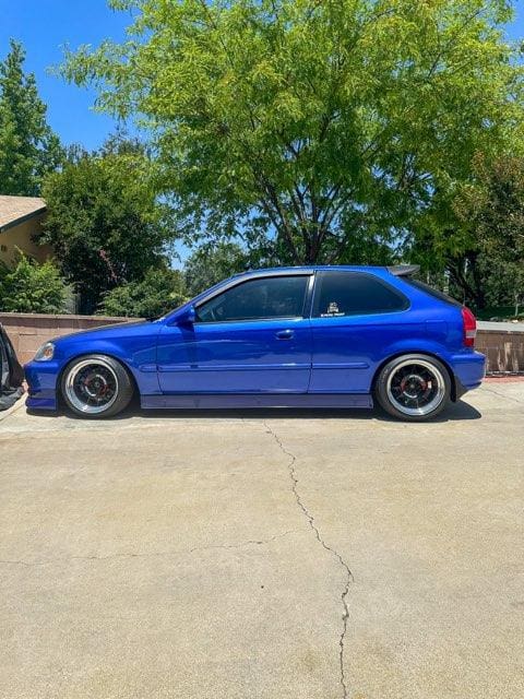 A blue hatchback car parked on a sunny street with greenery in the background.
