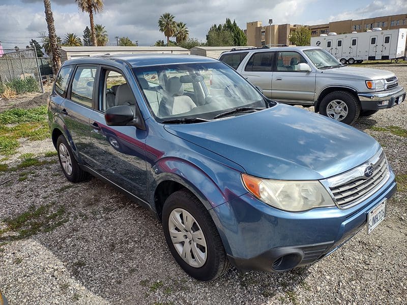 Blue compact SUV parked on gravel with palm trees in background.