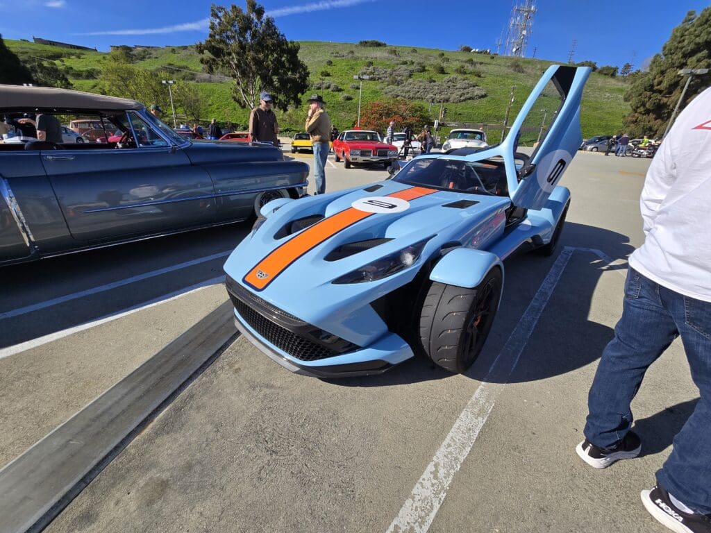 Blue sports car with orange racing stripes and gull-wing doors open in a parking lot.