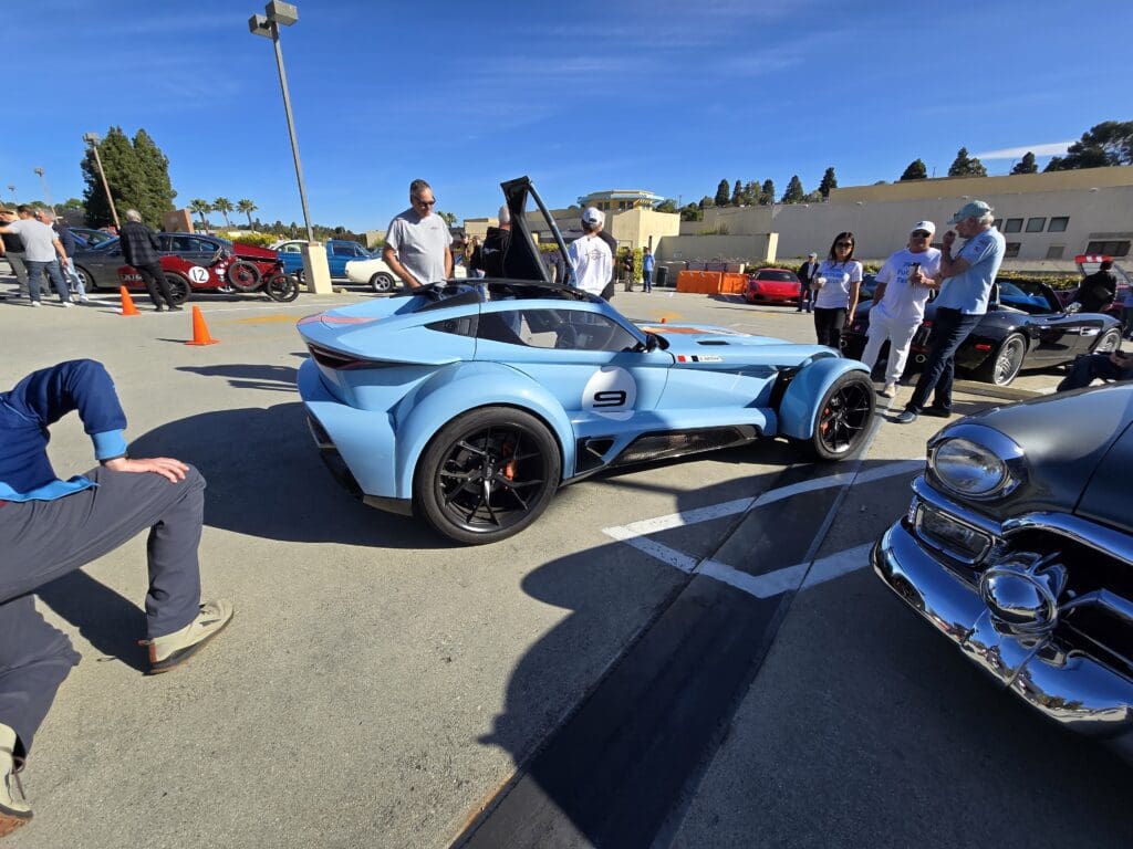 Blue sports car with orange racing stripes and gull-wing doors open in a parking lot.