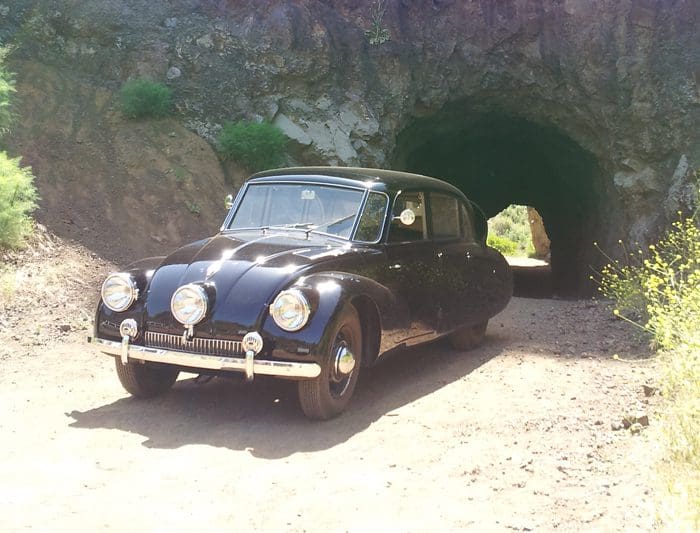 Vintage black car parked near a stone tunnel in a natural setting.