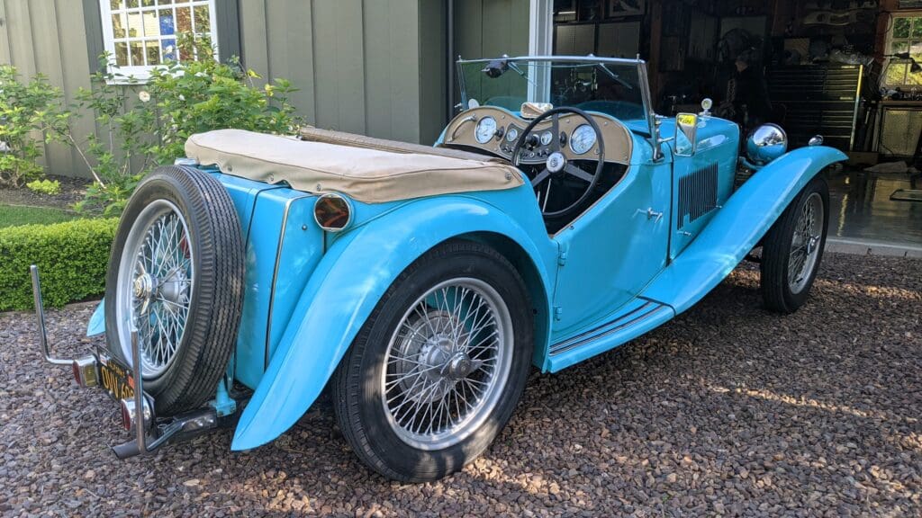 A vintage blue convertible car parked on gravel with wire-spoke wheels.
