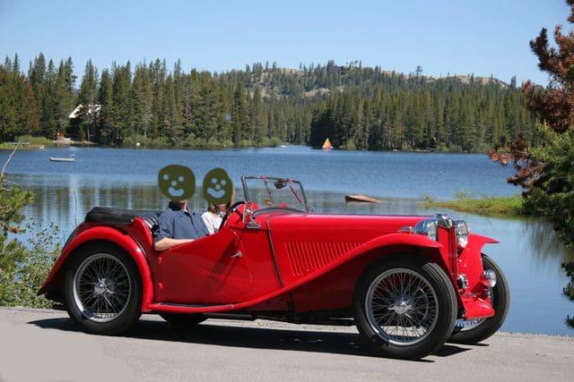 A classic red convertible car parked by a serene lakeside with forested hills in the background.