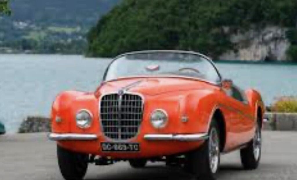 Vintage orange convertible car parked by a lake with forested hills in the background.