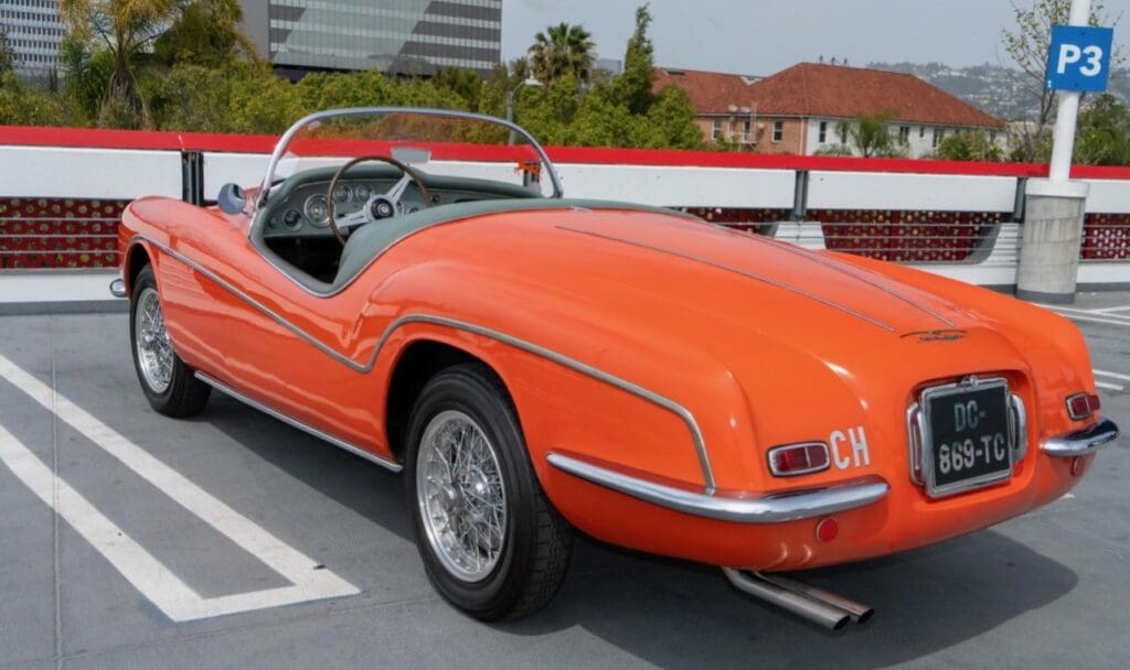Vintage orange convertible car parked by a lake with forested hills in the background.