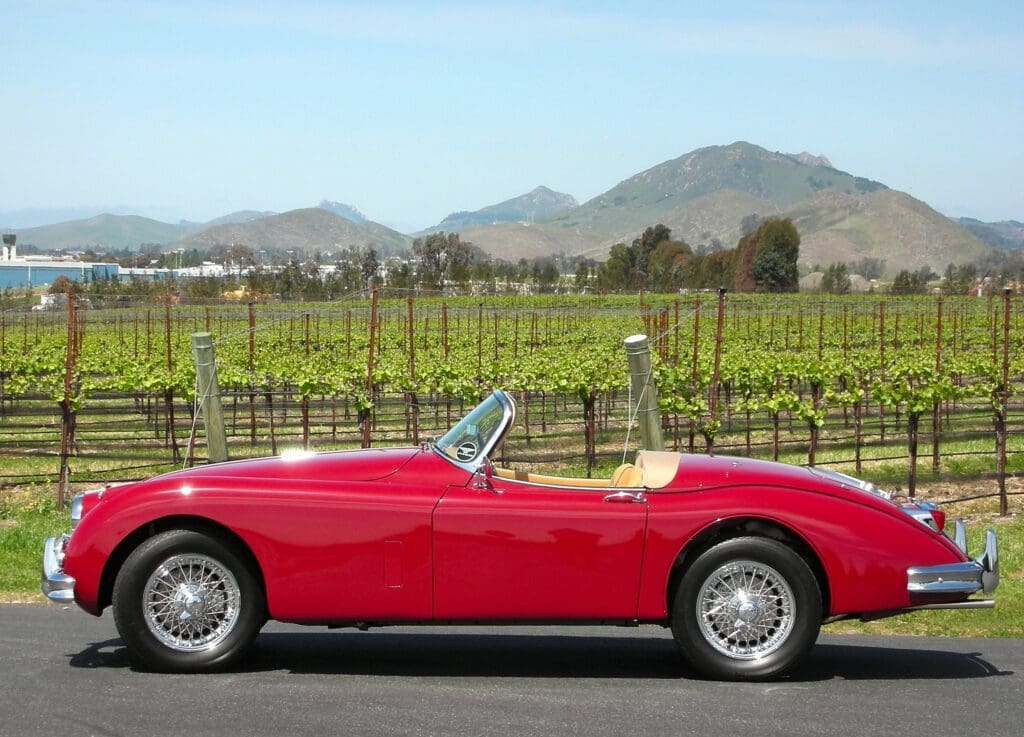 Classic red convertible sports car parked near a vineyard with mountains in the background.