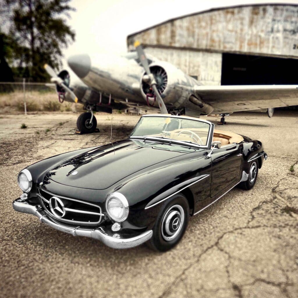Classic black convertible Mercedes parked near a vintage airplane hangar.