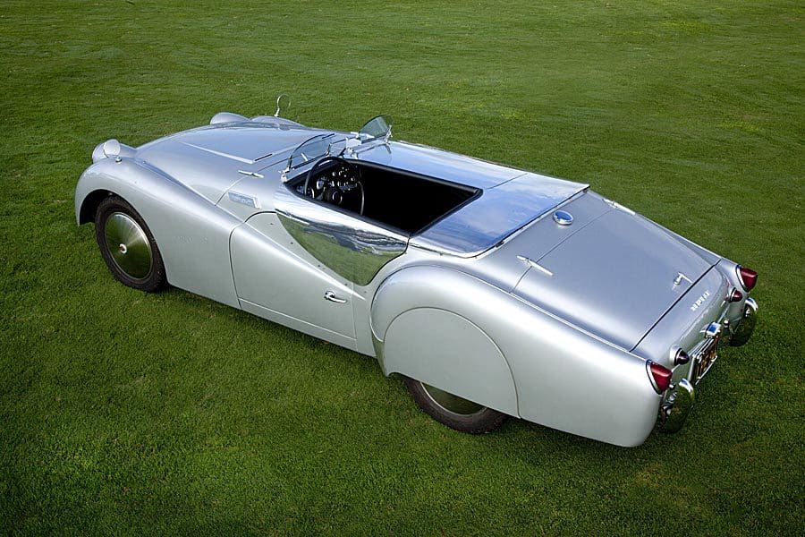 A sleek silver vintage car with a rear-mounted spare tire on a grassy field.