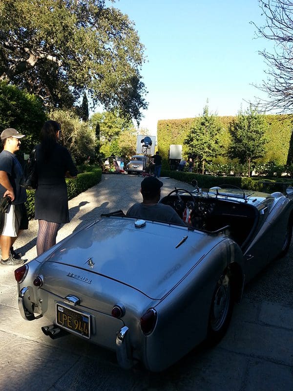A classic convertible car drives down a sunny suburban street with people nearby.