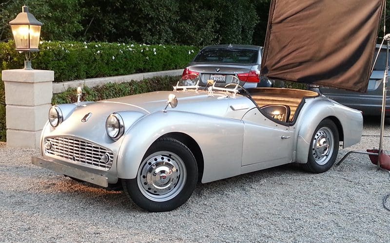Classic silver convertible car parked on gravel with greenery in background.