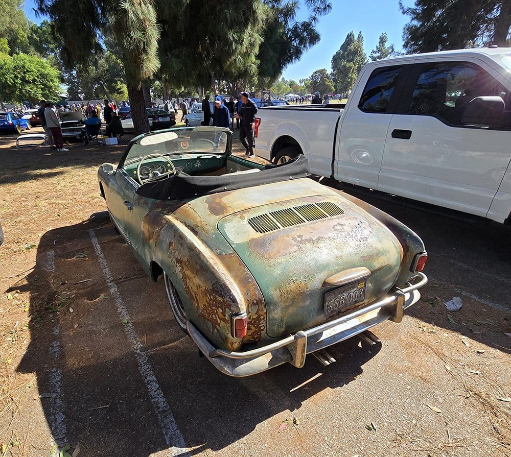 Rusty vintage car parked outdoors with a white truck nearby.