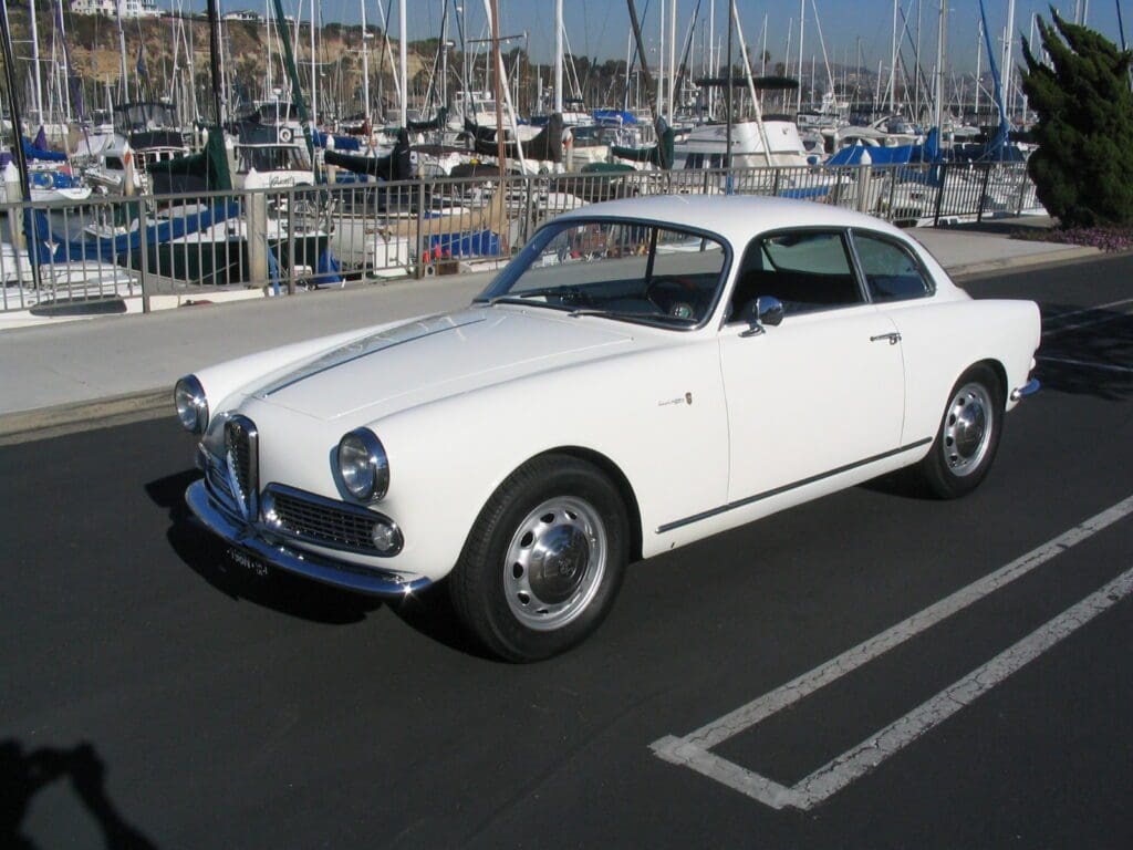 Classic white vintage car parked near a marina full of sailboats.