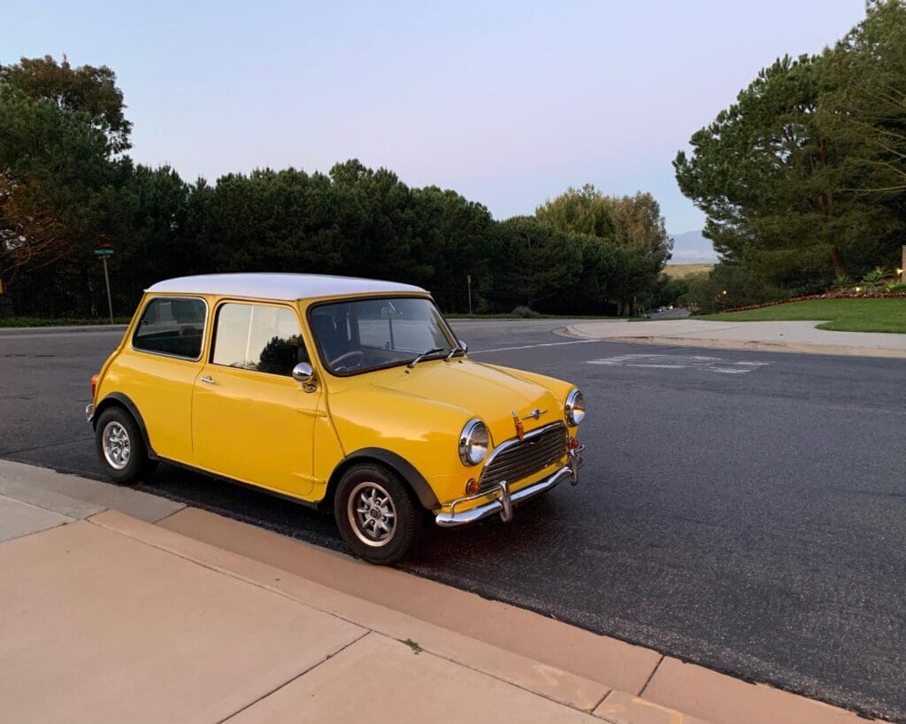 A classic yellow Mini Cooper parked by a roadside near greenery.
