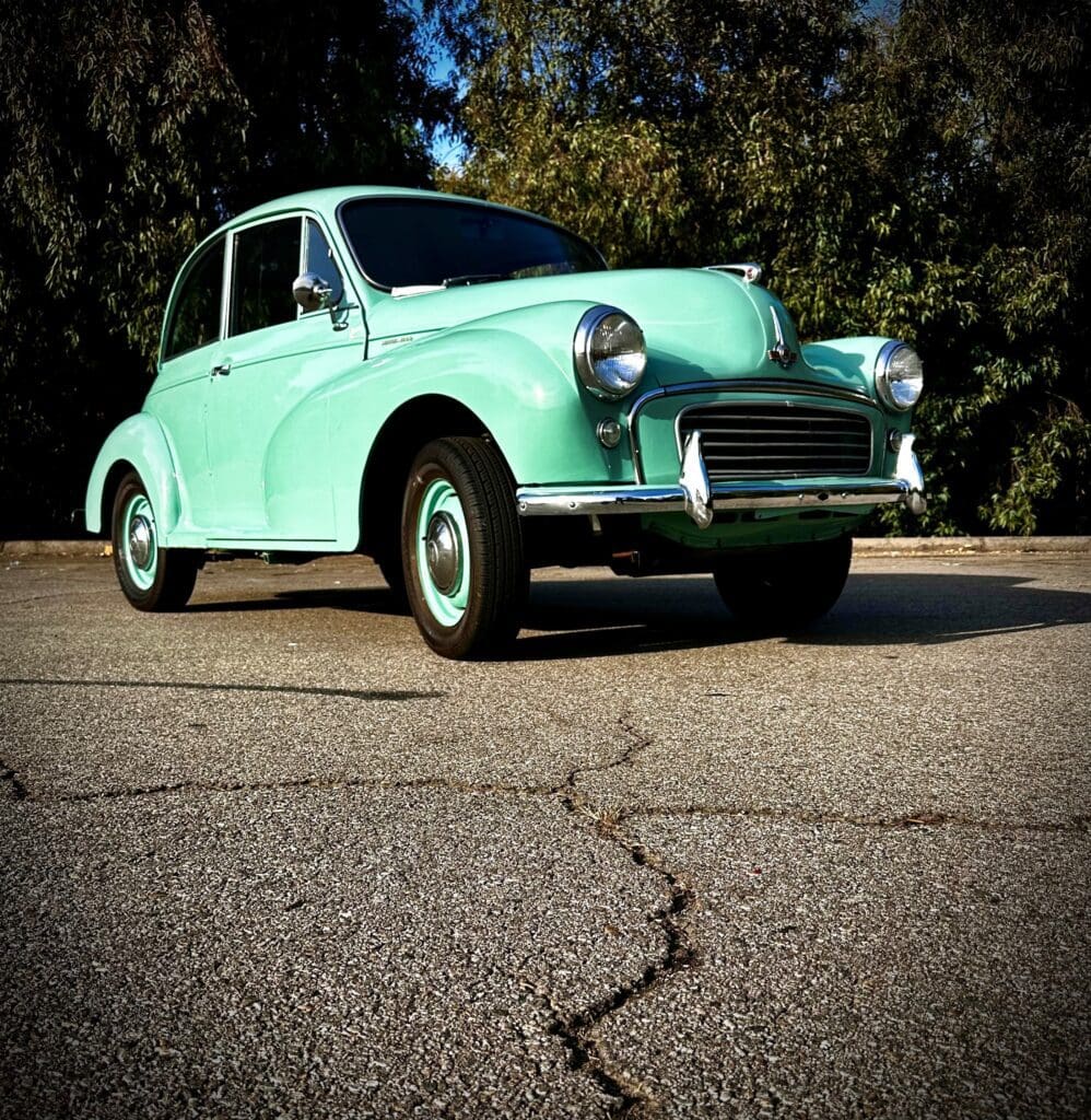 Light green vintage car parked on cracked pavement with trees in background.