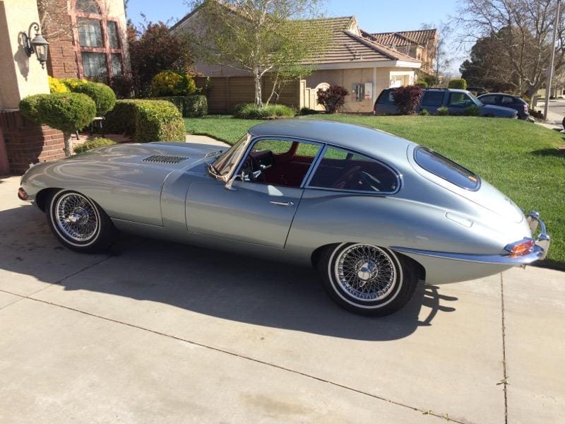 Classic silver Jaguar E-Type parked on a driveway in a sunny suburban neighborhood.