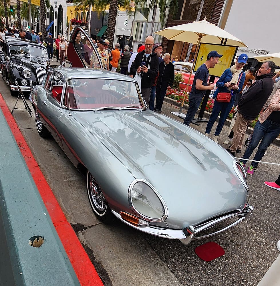 Classic silver sports car parked at a busy street event with onlookers.