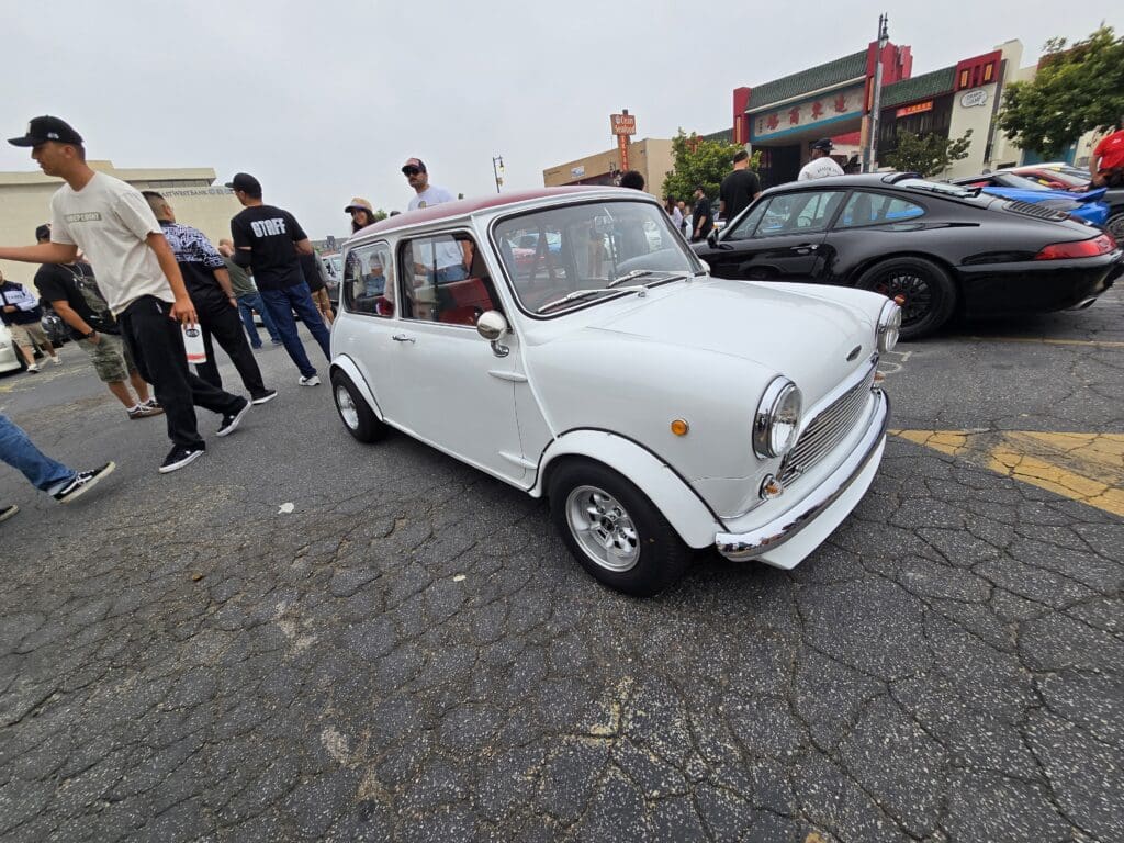A classic white Mini Cooper parked on a cobblestone street with onlookers around.