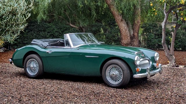 Classic green convertible car parked on gravel under trees.