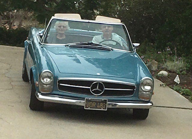 A classic blue Mercedes convertible with two passengers on a sunny day.