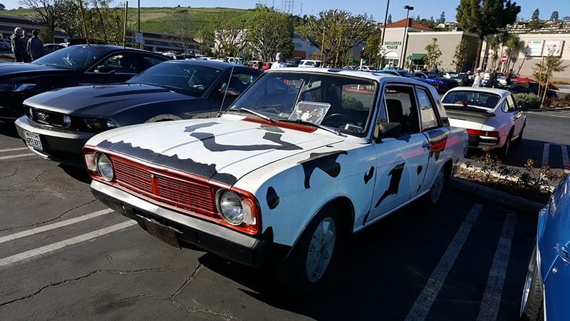A classic car painted with a cow pattern and horns on the hood.