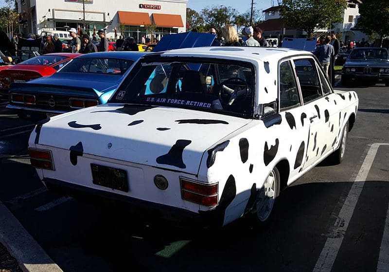 White car with black cow spots parked on street.