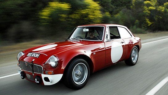 A classic red MG sports car with white racing decals driving on a road.