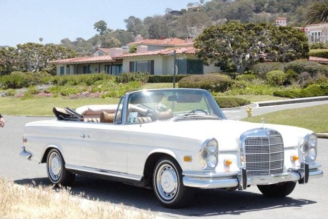 Classic white convertible car parked near a scenic coastal area with houses in the background.
