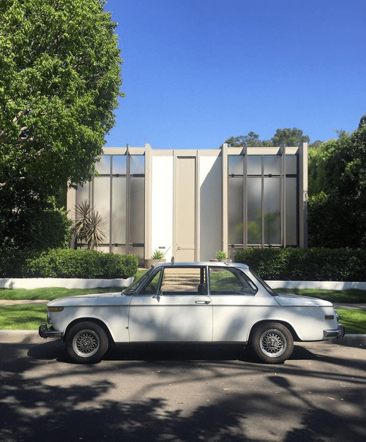 Classic white car parked in front of a modern house with large windows.
