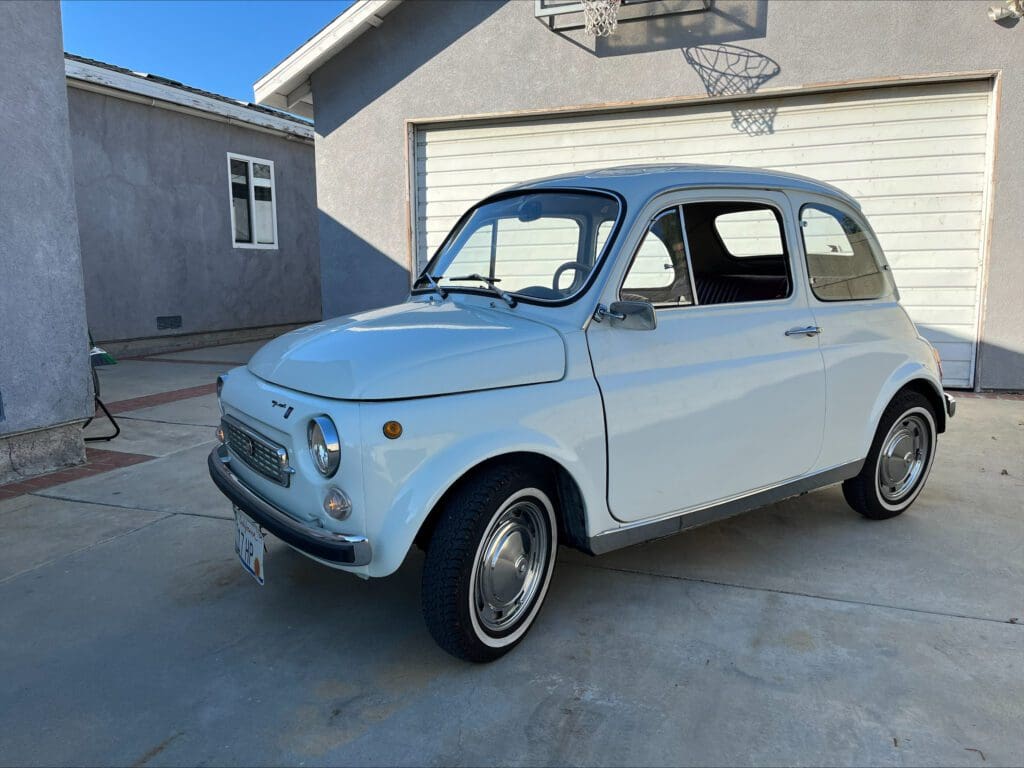 A vintage white compact car parked on a concrete driveway beside a building.