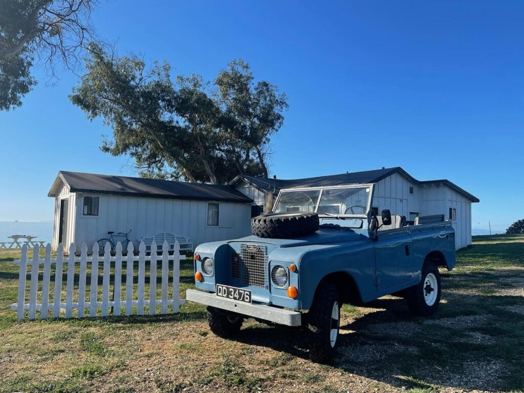 A vintage blue Land Rover parked beside a white picket fence under a clear blue sky.