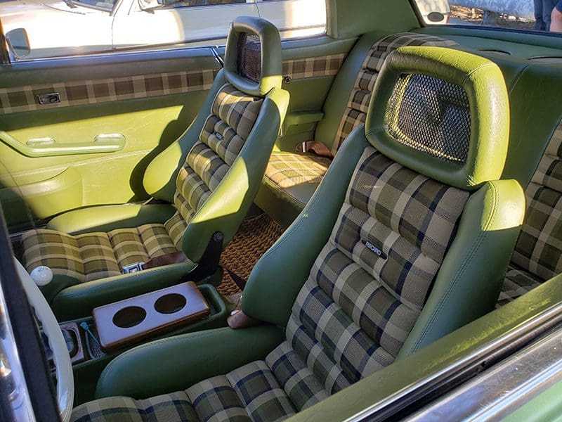 Vintage car interior with green plaid seats and center console cup holders.