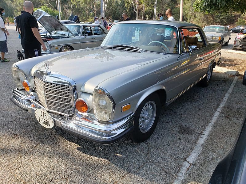 A classic silver Mercedes-Benz car parked outdoors with people nearby.
