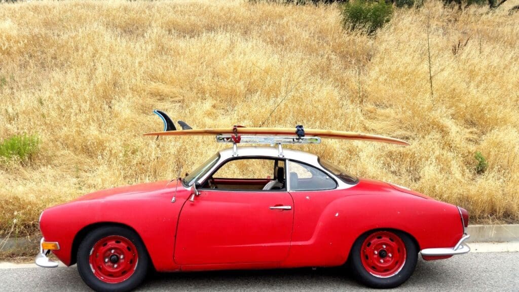 Red vintage car with surfboard on the roof parked in a dry grassy area.