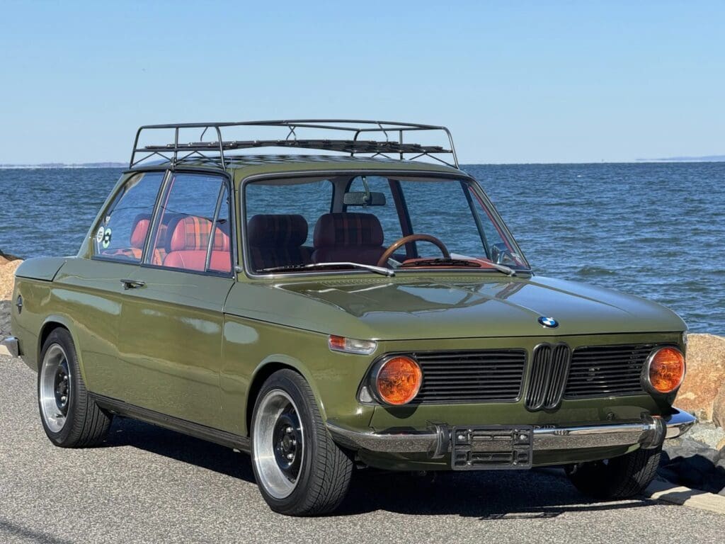 Classic olive-green vintage car parked by the sea under clear skies.