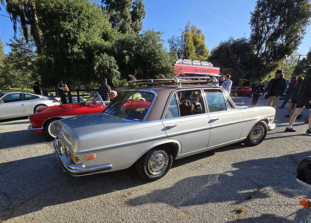 Classic silver car with roof luggage at a sunny outdoor event.