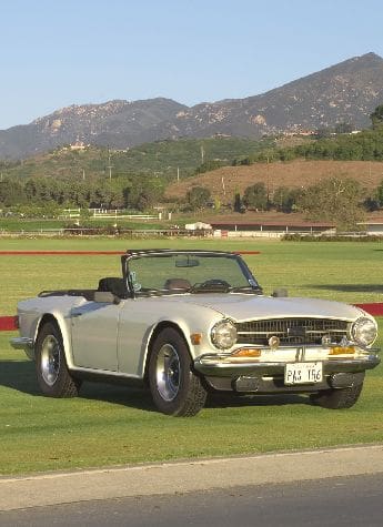 Classic silver convertible car parked on a grassy field with mountains in the background.