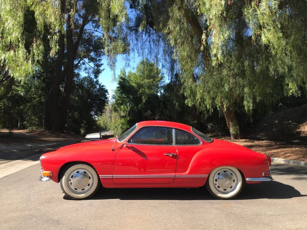 Classic red Volkswagen Karmann Ghia parked under large trees.