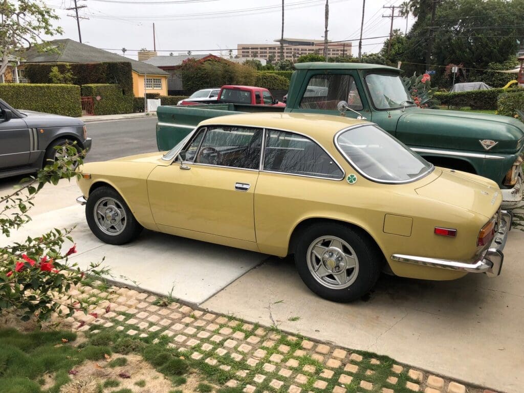 Classic yellow sports car parked on a driveway beside other vintage vehicles.