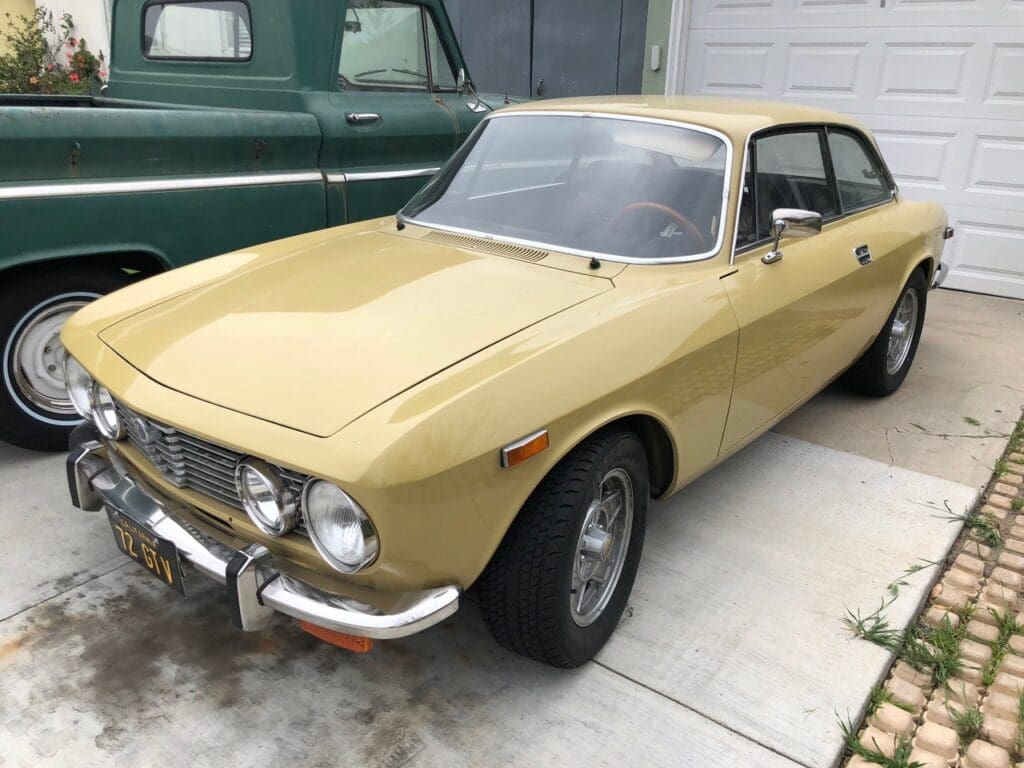 A vintage yellow car parked in a driveway next to a green vehicle.