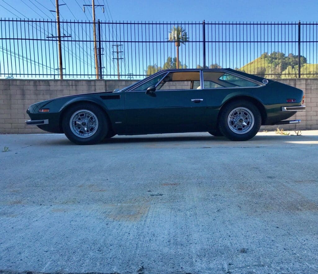 Classic dark green sports car parked beside a metal fence on a sunny day.